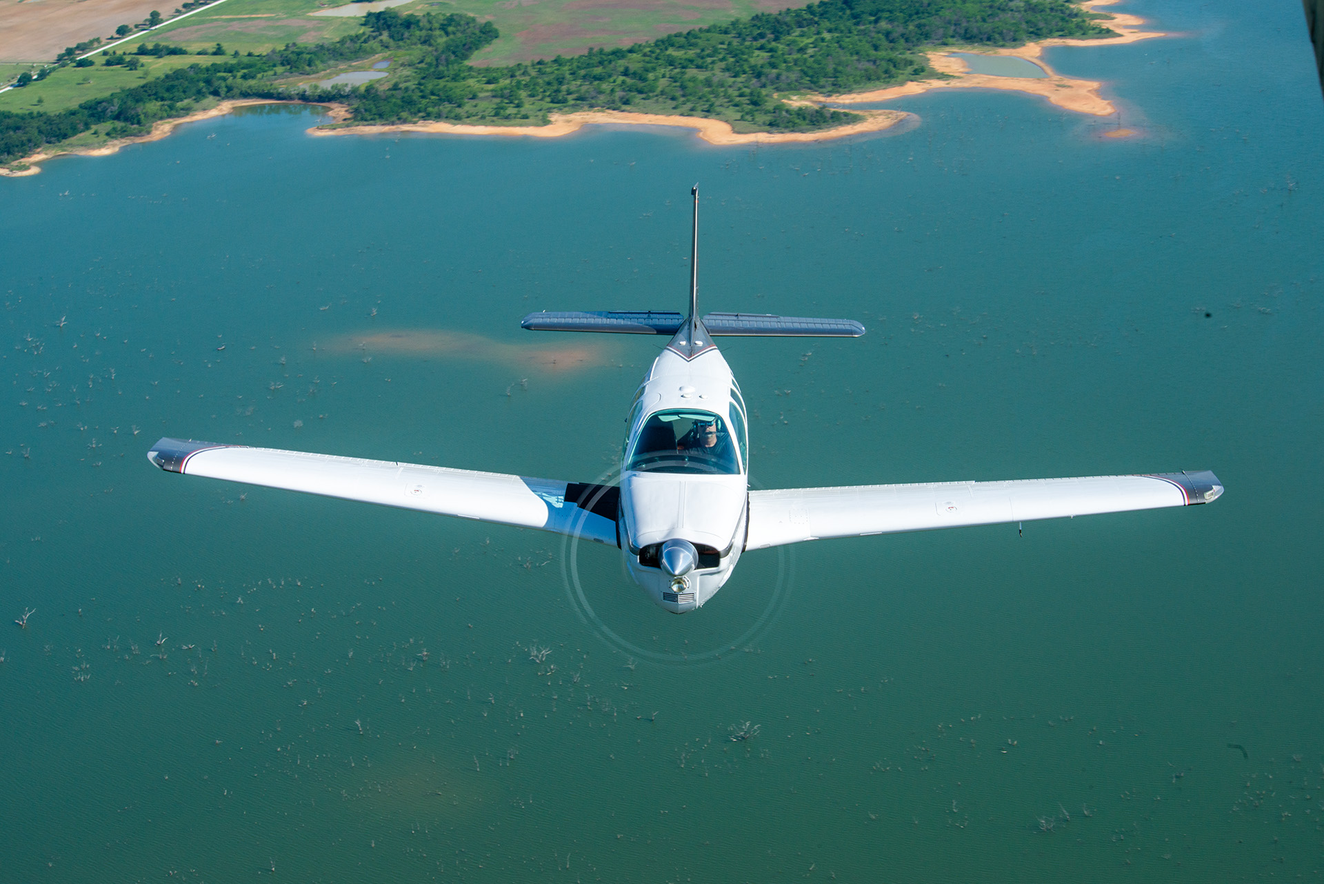 Aerial view of a single-engine aircraft flying over a body of water with a shoreline and green fields in the background.